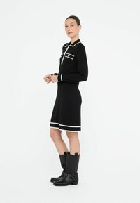Robe noire à manches longues et bordures blanches, avec un col, une fermeture éclair et des poches décoratives. Terminée avec des bottes noires montant jusqu'aux genoux.