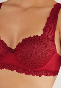 Czerwony koronkowy biustonosz z kwiatowymi wzorami, falbaniastymi krawędziami i gładkim materiałem, z fiszbinami i regulowanymi ramiączkami.