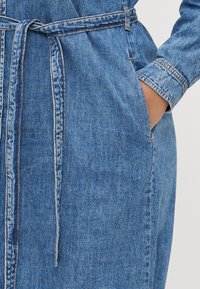 Denimkjole med belte i midjen, lange ermer og sidelommer. Mellomblå vask med synlig søm og en lett strukturert finish.
