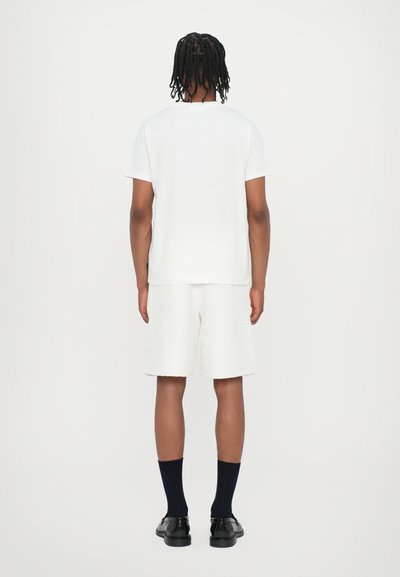 T-shirt bianca in cotone, abbinata a pantaloni corti di colore chiaro. Calzini e calzature neri completano l'outfit. Linee pulite e design semplice.