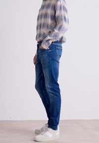 Jeans de denim azul con un corte slim, que cuentan con cinco bolsillos y costuras sutiles. Combinados con una camisa de cuadros clara y zapatillas blancas.