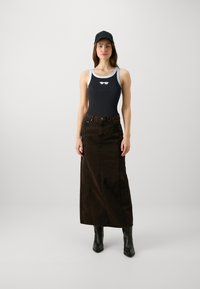 En ung kvinna som bär en svart keps, svart linne med solglasögonlogotyp, lång mörkbrun denimkjol och svarta anklarstövlar, står rak.