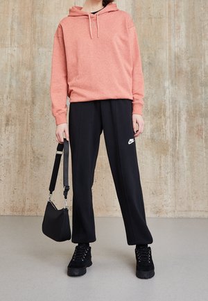 Personne portant un sweat à capuche rose et un pantalon noir Nike, tenant un sac à main noir, debout contre un mur texturé beige.