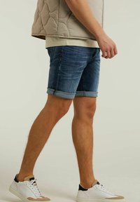 Hombre que lleva unos pantalones cortos de denim arremangados, un chaleco acolchado beige, una camisa clara y zapatillas blancas con acentos en marrón y negro, de pie contra un fondo plano.