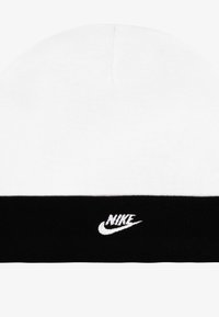 Vit stickad mössa med en svart ribbad kant, som har en liten vit Nike-logotyp broderad på kanten. Mjuk textur, rundad form.