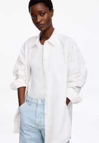 Femme aux cheveux courts portant une chemise blanche boutonnée sur un débardeur blanc et un jean bleu clair, mains dans les poches, sur un fond uni.
