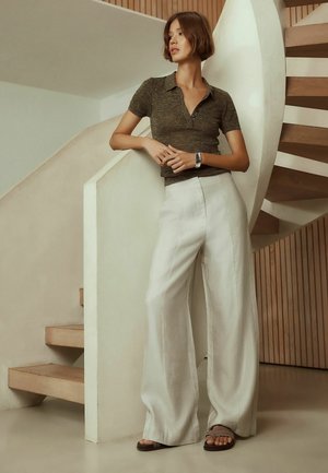 Tânără îmbrăcată într-un top de culoare închisă cu mâneci scurte și pantaloni largi albi se sprijină de un perete alb curbat lângă o scară spirală de lemn în interior.