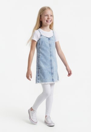 Mladé dievča sa usmieva, kráča, má na sebe svetlomodré rifľové šaty cez biele tričko a biele legíny s bielymi teniskami.