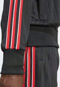 Černá sportovní souprava svislými proužky, zvýrazněná červenými a bílými pruhy na rukávech a kalhotách. Texturovaná látka.