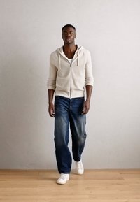 Lys grå hettegenser med glidelås over en hvit skjorte, kombinert med mørkeblå jeans og hvite joggesko. Modellen står på tregulv mot en nøytral vegg.
