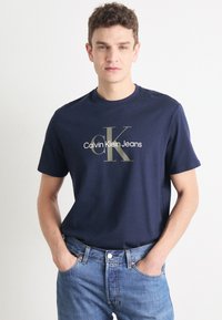 Camiseta de algodón azul marino con cuello redondo y mangas cortas, que presenta un logo impreso en dorado "Calvin Klein Jeans" en el frente.