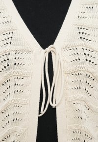 Πλεκτή ζακέτα σε κρεμ απόχρωση με V-λαιμόκοψη, ανοιχτό μπροστινό μέρος με δέσιμο και υφασμένο δαντελένιο σχέδιο στους πάγκους.