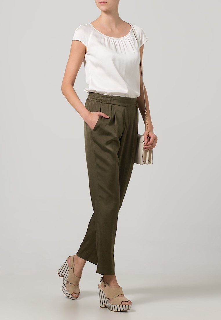 Femme portant un chemisier blanc à manches courtes, un pantalon taille haute vert olive, des talons compensés beiges, et tenant un petit sac blanc avec une chaîne dorée.