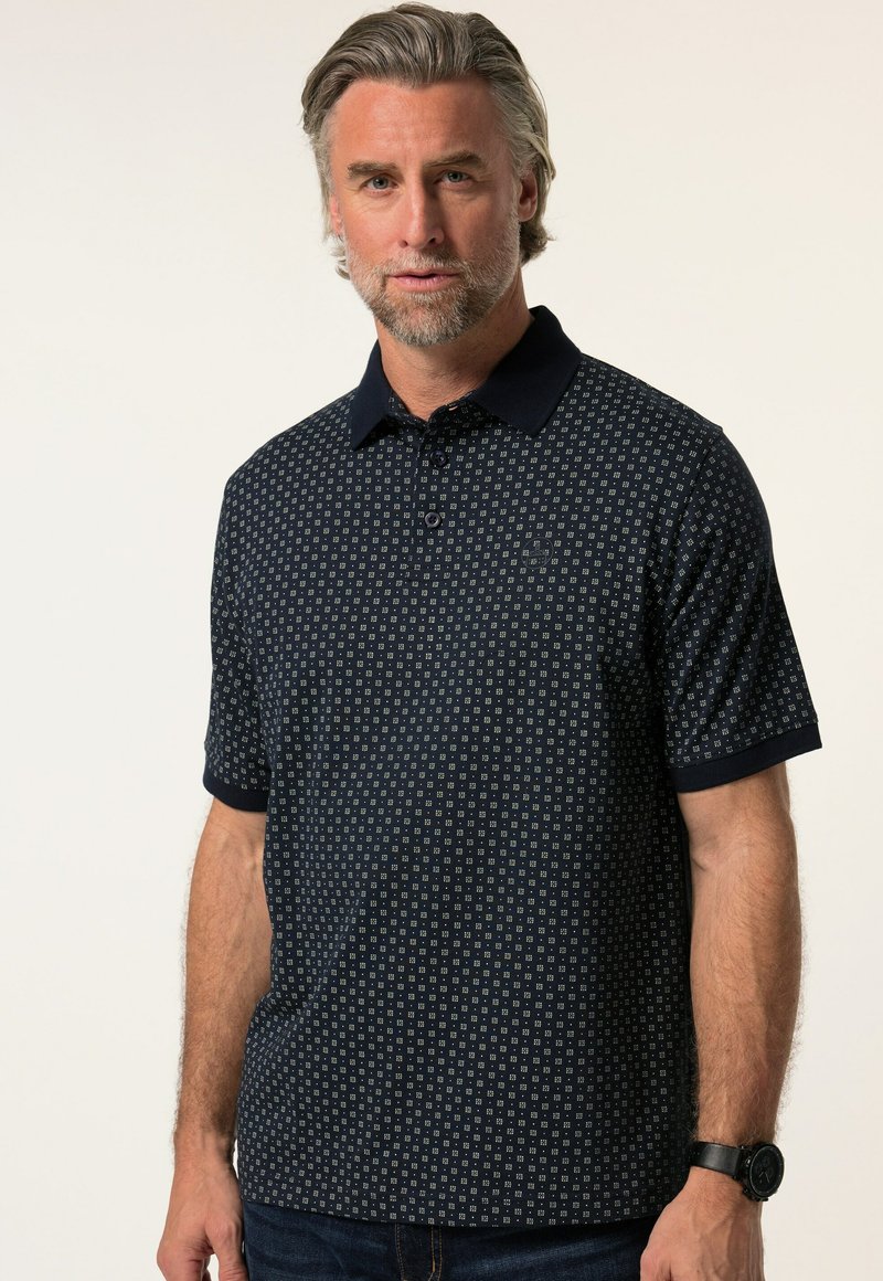 Tummansininen polo-paita lyhyillä hihoilla, jossa on geometrinen kuvio ja kontrastivärinen kaulus. Pehmeä pinta, jossa nappilista yksityiskohtana.