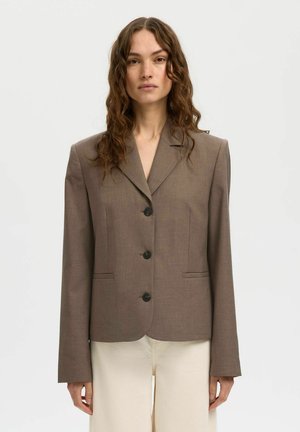 Ženska z valovitimi rjavimi lasmi oblečena v krojen rjav blazer s tremi črnimi gumbi in krem hlače na enobarvnem ozadju.