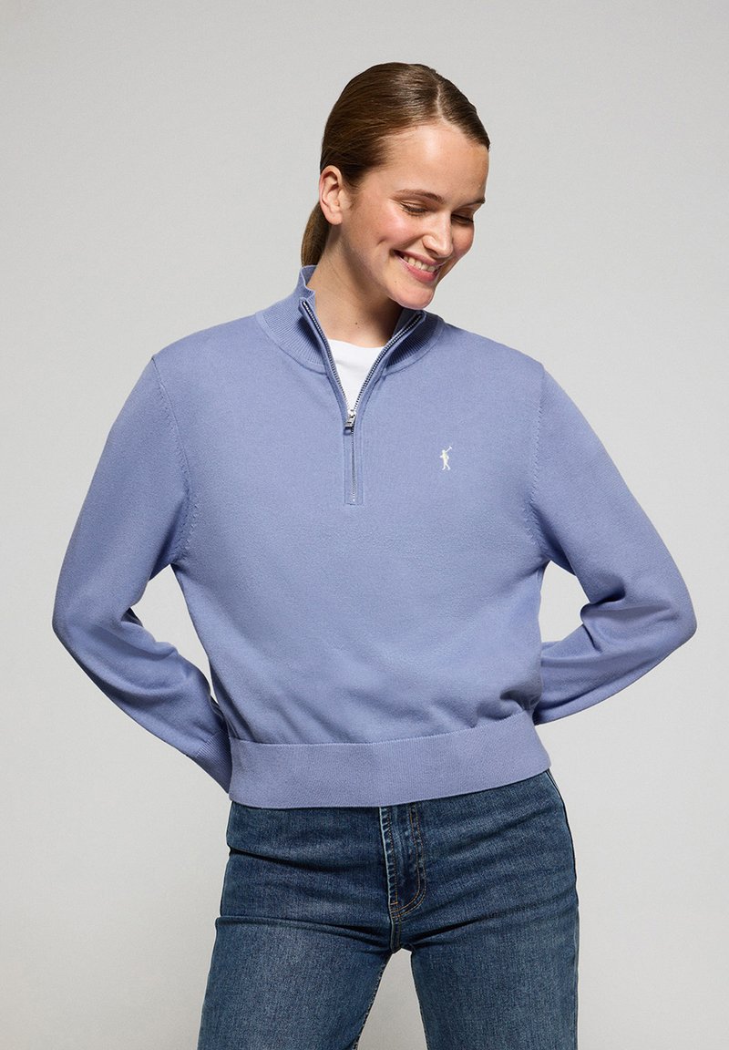 Ljusblå tröja med dragkedja i kragen, ribbade manschetter och avslut, med en liten vit logotyp på vänster bröst. Bärs med blå jeans.