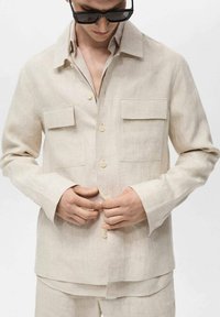 Bež srajca iz lana s sproščenim dizajnom, ki vključuje dva žepa na prsih, zaklepanje z gumbi in dolge rokave. Gladka textura, lahka tkanina.