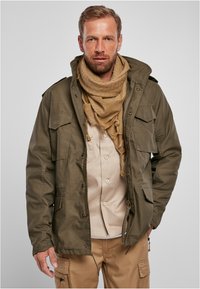 Chaqueta verde oliva con capucha y múltiples bolsillos frontales, llevada sobre una camisa beige. Completada con una bufanda marrón, que presenta un patrón texturizado.