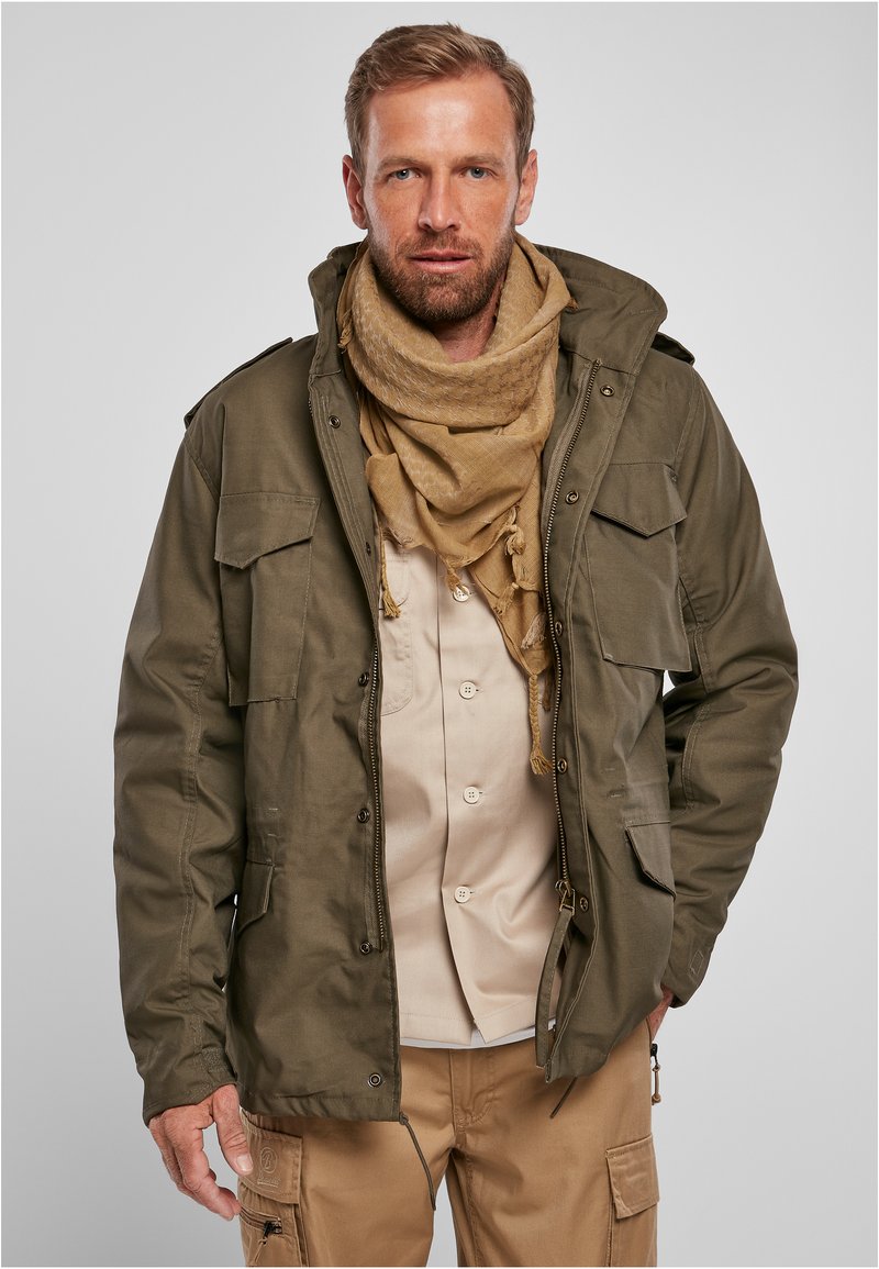 Chaqueta verde oliva con capucha y múltiples bolsillos frontales, llevada sobre una camisa beige. Completada con una bufanda marrón, que presenta un patrón texturizado.