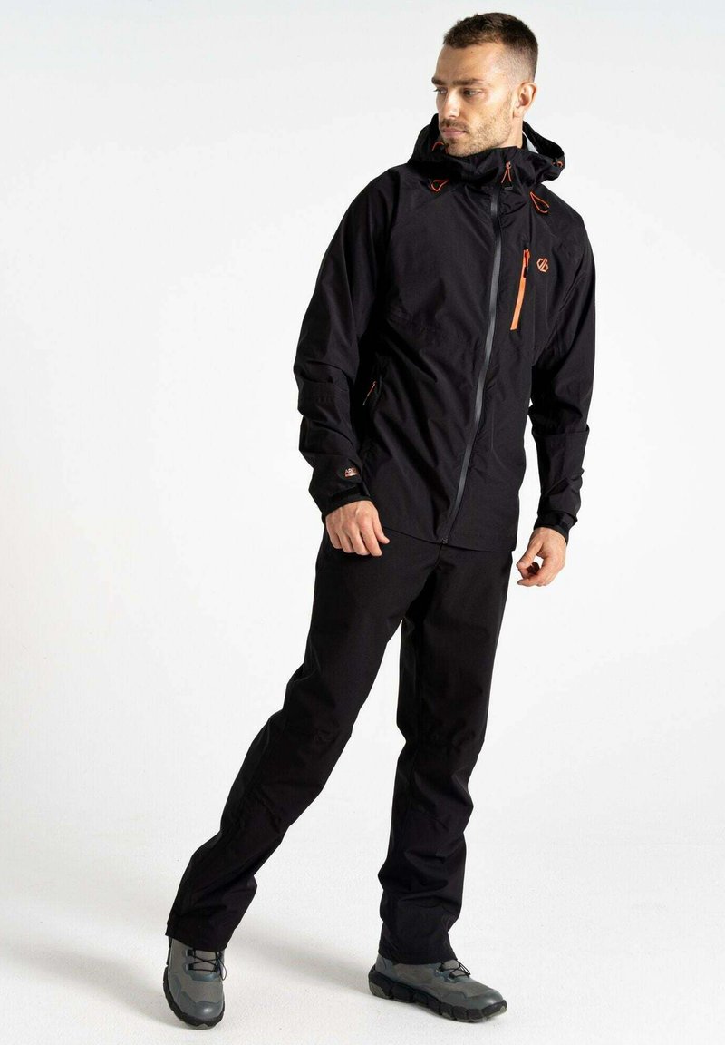 Giacca impermeabile nera con accenti grigi, cerniere arancioni, cappuccio regolabile e tasche con zip, indossata con pantaloni neri coordinati e scarpe grigie.