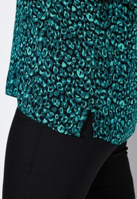 Top a maniche corte in teal e nero con un motivo leopardato, caratterizzato da spacchi laterali e una vestibilità rilassata, abbinato a leggings neri.