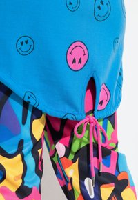 Top azul brillante con estampados de caritas sonrientes, con una abertura lateral y cordón rosa. Leggings coloridos con un patrón abstracto completan el atuendo.