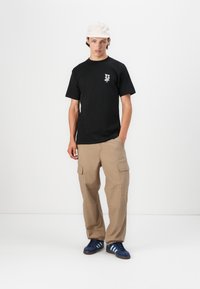 Sort bomuld t-shirt med et lille grafisk design på venstre bryst, parret med beige cargo bukser med to sidelommer og marineblå sneakers.