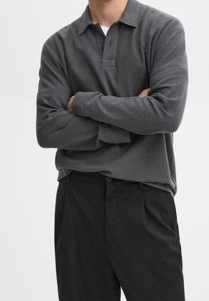 Homme portant un polo à manches longues gris foncé avec col boutonné et pantalon noir, les bras croisés sur la poitrine devant un fond uni.