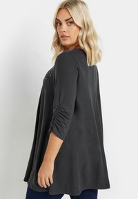 Donkere, grijze top met lange mouwen en een losse pasvorm, voorzien van gathered mouwen en versieringen in een subtiel patroon over de rug.