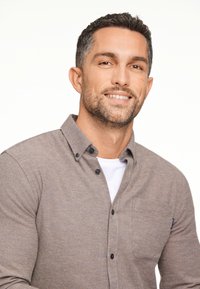 Camicia marrone a bottoni con taschino, indossata sopra una T-shirt bianca. Il tessuto sembra morbido, con una finitura texture e bottoni scuri.