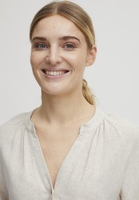 Svetlo béžová ľanová blúzka s okrúhlym výstrihom, jemnými nariaseniami a voľným strihom. Hladká textúra s prirodzeným, vzdušným vzhľadom.