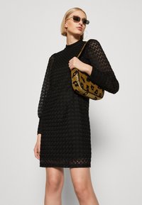 Vestido preto com tecido texturizado e padronizado; mangas longas e decote alto. Modelo segura uma bolsa dourada com padrão de leopardo. Óculos de sol estilosos completam o visual.