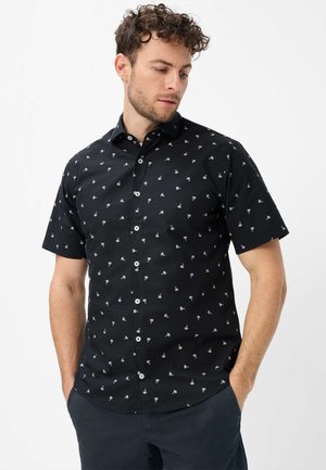 Mann mit lockigem Haar trägt ein schwarzes kurzärmeliges Hemd mit kleinen weißen Palmenmotiven und dunkle Hosen, Hände in den Taschen, schaut nach unten.