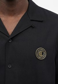 Άνδρας που φοράει μαύρο πουκάμισο με κουμπιά και χρυσή και μαύρη κυκλική ρεζέρβα λογότυπου Just Cavalli στο αριστερό στήθος.