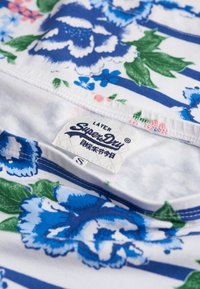Blumenmusterstoff mit blauen und grünen Blumen auf weißem Hintergrund, mit horizontalen blauen Streifen. Enthält ein "SuperDry"-Label.