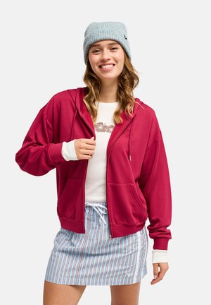 Junge Frau mit grauer Strickmütze, rotem Kapuzenpullover mit Reißverschluss, weißem Langarmshirt und blau gestreiftem Rock, lächelnd mit einer Hand, die ihre Jacke anpasst.