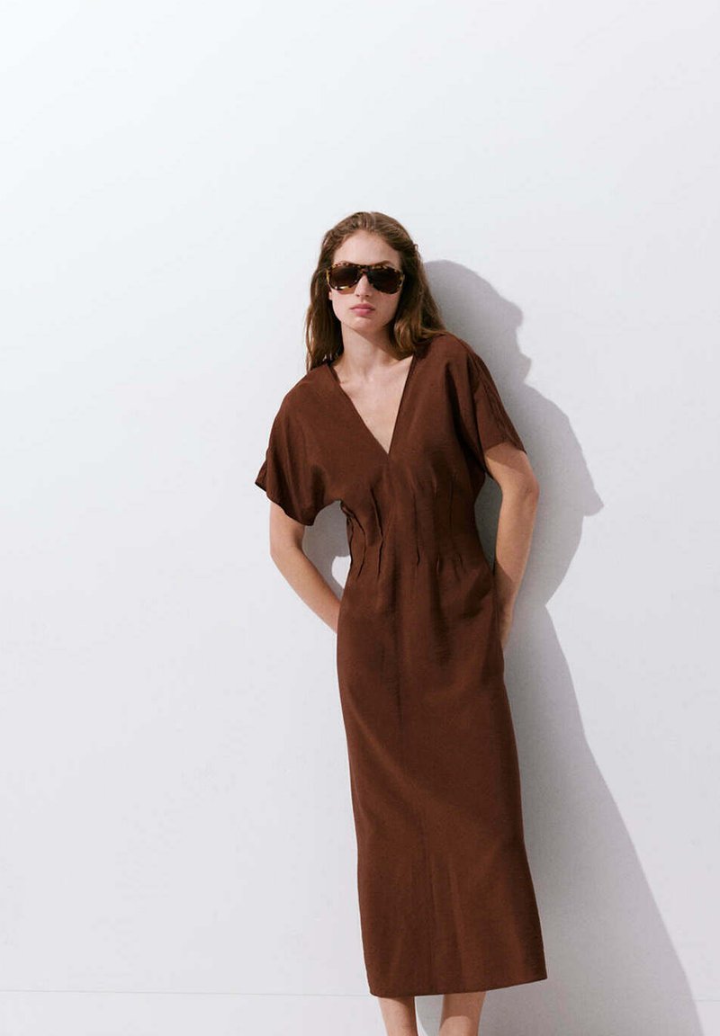 Femme portant une robe midi marron à col en V et de grandes lunettes de soleil, debout contre un mur blanc, les mains dans les poches.