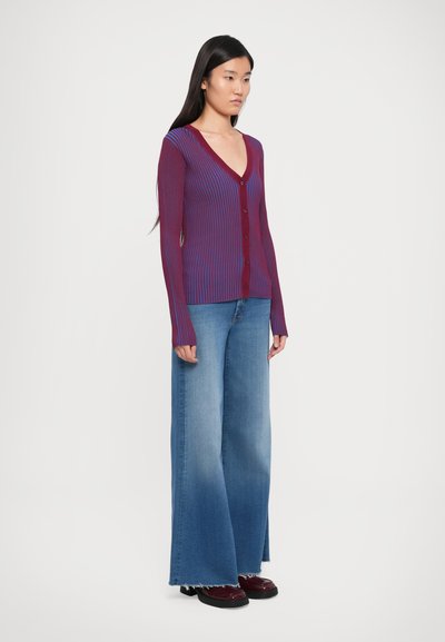 Rød og blå stripete ribbestrikket cardigan med V-hals, knappestolpe foran og lange ermer. Kombinert med vide lyseblå jeans og mørke sko.