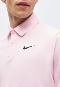 Homme portant un polo rose clair avec le logo noir Nike swoosh, col visible et deux boutons fermés, visage partiellement visible.