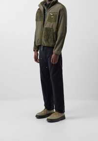 Bluza polarowa w oliwkowej zieleni z teksturowanym panelem i kieszenią na suwak; zestawiona z ciemnymi spodniami i beżowymi butami z żółtymi sznurowadłami.