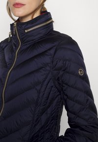 Kvinna klädd i en marinblå quiltad jacka med guldfärgad dragkedja och Michael Kors-logotyp på ärmen, stående mot en vit bakgrund.