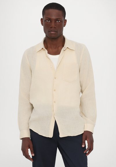 Camicia di lino beige con maniche lunghe, bottoni frontali, taschino sul petto singolo e una vestibilità leggermente rilassata, indossata sopra una canottiera bianca.