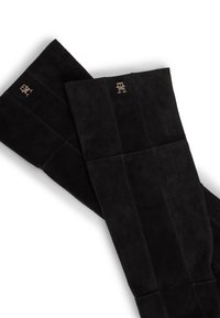 Bottes en suède noir au-dessus du genou, présentant une texture lisse, des coutures verticales et des accents de logo en argent signature en haut.