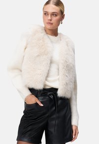 Weiße Kunstfell-Jacke in Cropped-Form über einem enganliegenden wei�ßen Sweater, kombiniert mit hoch taillierten schwarzen Leder-Shorts mit gebundenem Bund und seitlichen Taschen.