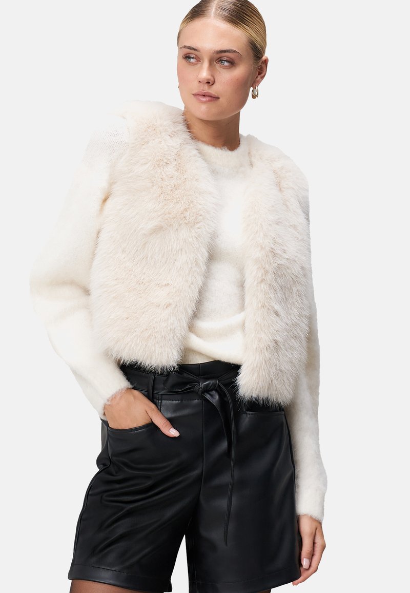 Weiße Kunstfell-Jacke in Cropped-Form über einem enganliegenden weißen Sweater, kombiniert mit hoch taillierten schwarzen Leder-Shorts mit gebundenem Bund und seitlichen Taschen.