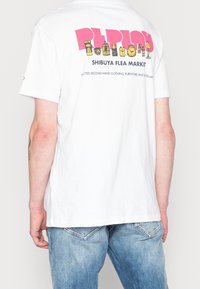 Hvid bomulds T-shirt med et farverigt grafisk tryk på ryggen, der viser "SHIBUYA FLEA MARKET" med forskellige ikoner.