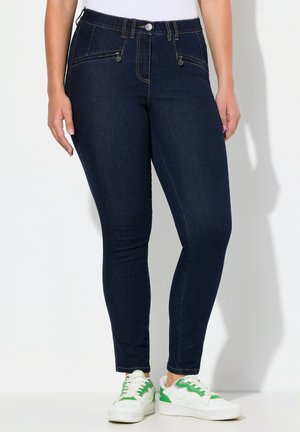 Frau trägt dunkelblaue Skinny-Jeans mit vorderen Reißverschlusstaschen und weiße Sneaker mit grünen Akzenten, steht vor einem weißen Hintergrund.