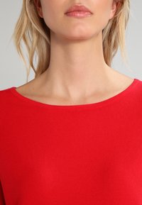 Pull rouge côtelé avec un décolleté large et arrondi. Le tissu a une texture lisse, montrant des détails de couture subtils.