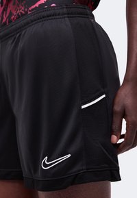 Μαύρα σορτς Nike με λευκό λογότυπο Swoosh, φορεμένα από άτομο με σκουρόχρωμο δέρμα, με πλεγμένες πλαϊνές επενδύσεις και ελαστική μέση.