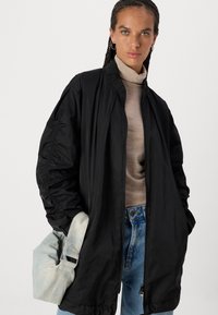 Schwarze Bomberjacke mit Reißverschluss, elastischen Bündchen und seitlichen Taschen, kombiniert mit einem beigen Rollkragenpullover und hellblauen Jeans.